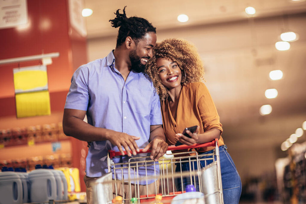 African American couple choosing products using phone during grocery shopping in modern supermarket - Zdjęcie, obraz