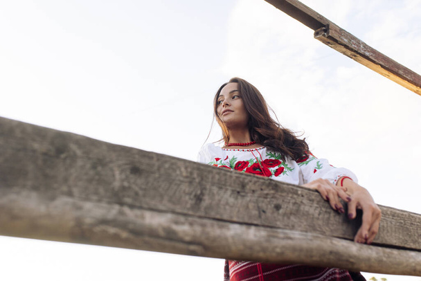 Young Ukrainian woman stands in traditional national embroidered shirt and skirt near wooden fence against sky background. Bottom view. Ethnic ukrainian national clothes style, embroidered shirt. - Fotó, kép