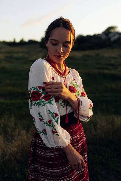 Молода українка стоїть у традиційній національній вишитій сорочці та спідниці на лузі під час заходу сонця. Етнічний національний стиль одягу, вишита сорочка.. - Фото, зображення