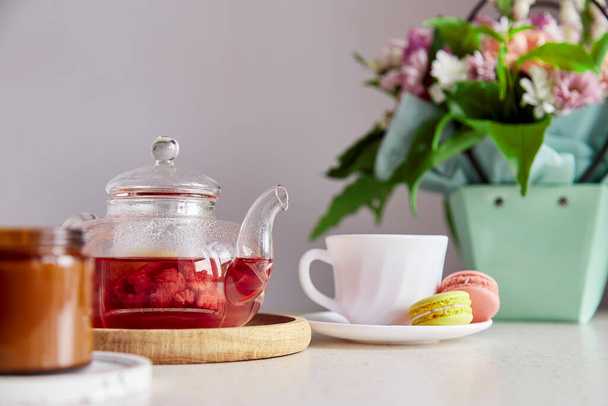 Autumn tea time - cup of tea with macarons, glass teapot, candle and bouquet of flowers. Herbal raspberry natural tea. Cozy home with tea and dessert. Self care, relaxing, wellness lifestyle - Fotoğraf, Görsel