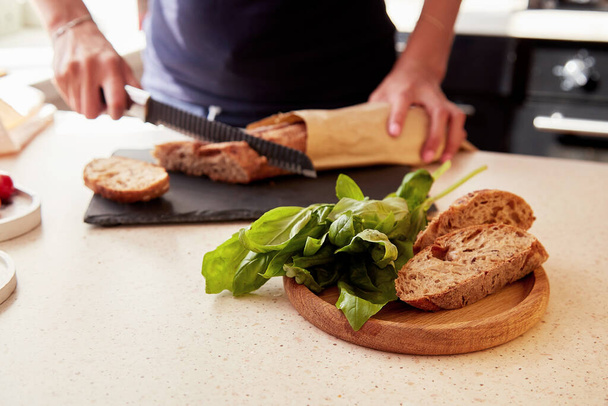 Woman hands is cutting a French baguette in the kitchen. Basil and whole grain cutting bread. Selective focus. Blurred background. - Fotografie, Obrázek