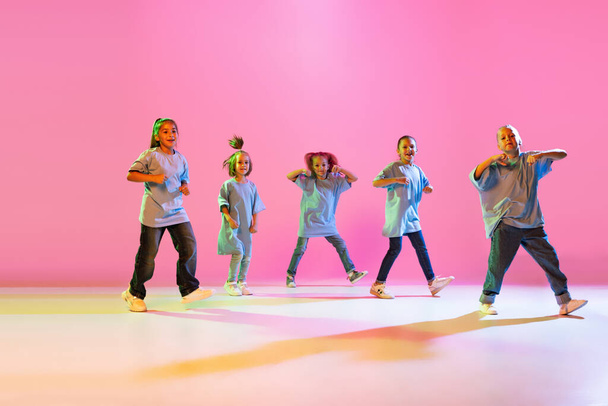 Hip-hop dance, street style. Happy children, little active girls in casual style clothes dancing isolated on orange background in purple neon light. Concept of music, fashion, art - Fotografie, Obrázek