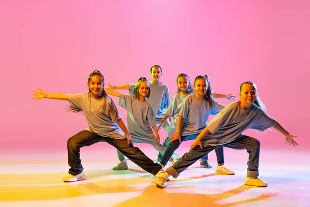 Hip-hop dance, street style. Happy children, little active girls in casual style clothes dancing isolated on orange background in purple neon light. Concept of music, fashion, art - Valokuva, kuva