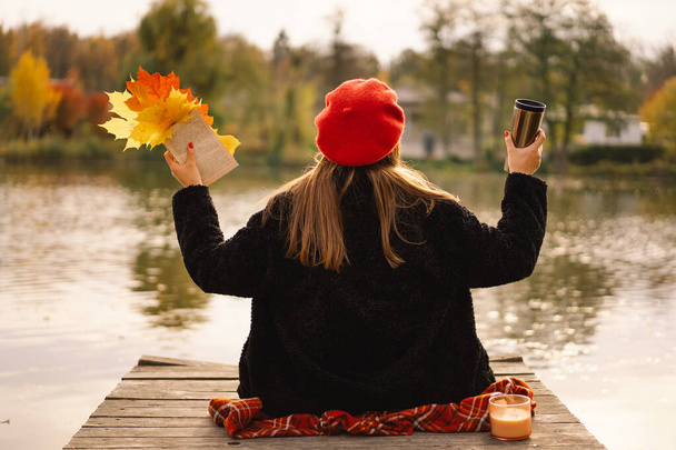 Woman in a red beret reading book on wooden pontoon. Autumn season. Female with trendy clothes relaxing near water reading book and drink coffee. Autumn mood. Autumn concept. Autumn day. - Fotó, kép