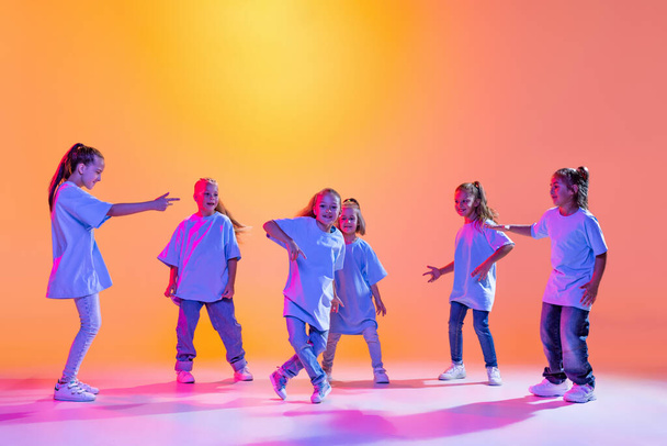 Hip-hop dance, street style. Happy children, little active girls in casual style clothes dancing isolated on orange background in purple neon light. Concept of music, fashion, art - Фото, изображение