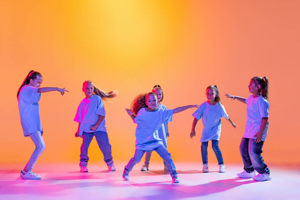 Happy children dancing. Group of children, little girls in sportive casual style clothes dancing in choreography class isolated on orange background in purple neon light. Concept of music, fashion - Фото, изображение