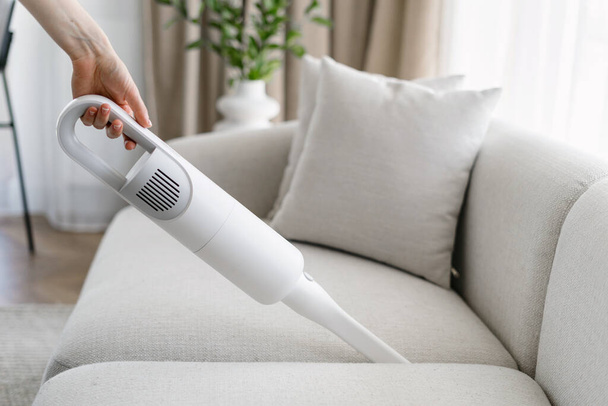 Side view of woman cleaning soft sofa using cordless vacuum in living room. Concept of cleanliness. Household equipment usage. Housewife chores. Cleaning service worker duties - Fotó, kép