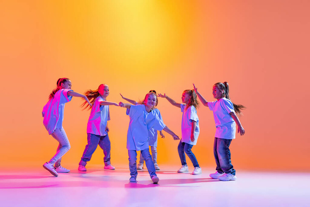 Modern choreography lesson. Dance group of happy, active little girls in t-shirts and jeans in action isolated on orange background in neon. Concept of music, fashion, art, childhood, hobby. - Foto, Bild