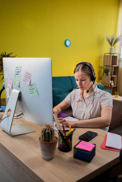 Portrait view of focused young woman in headset with microphone sitting on sofa, involved in video call conversation. Female having online meeting workshop with colleagues. - Fotografie, Obrázek