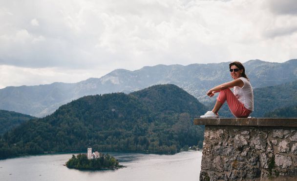 Young smiling woman tourist sitting on a Bled Castle wall with the lake Bled and the Bled island on the background. Surrounded by mountains and forests in Slovenia. Travel concept - Foto, imagen