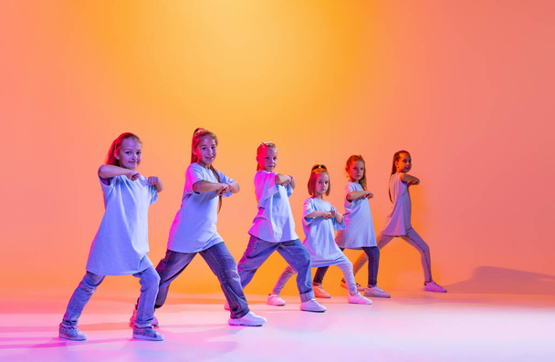 Joy, fun and happiness. Portrait of cheerful, active little girls, happy kids dancing isolated on orange background in neon light. Concept of music, fashion, art, childhood, hobby. - Foto, Imagem