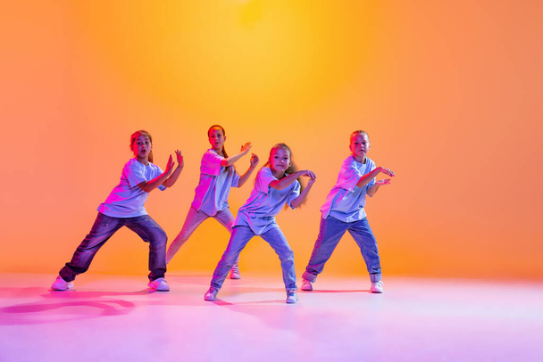 Happy children dancing. Group of children, little girls in sportive casual style clothes dancing in choreography class isolated on orange background in purple neon light. Concept of music, fashion - Foto, Imagem