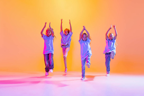 Modern choreography lesson. Dance group of happy, active little girls in t-shirts and jeans in action isolated on orange background in neon. Concept of music, fashion, art, childhood, hobby. - Photo, Image