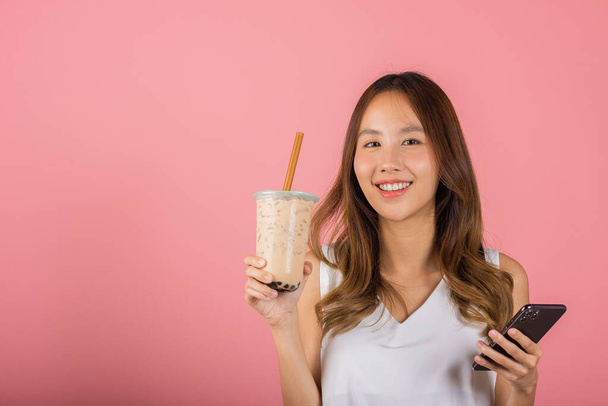 Asian beautiful young woman holding drinking brown sugar flavored tapioca pearl bubble milk tea, Smiling portrait female hold smartphone, studio shot isolated on pink background, milk beverage concept - Foto, Imagem