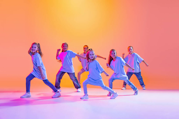 Hip-hop dance, street style. Happy children, little active girls in casual style clothes dancing isolated on orange background in purple neon light. Concept of music, fashion, art - 写真・画像