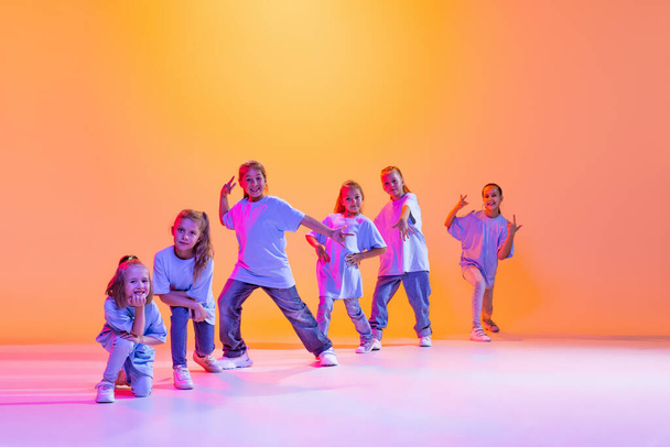 Happy children dancing. Group of children, little girls in sportive casual style clothes dancing in choreography class isolated on orange background in purple neon light. Concept of music, fashion - Foto, Bild