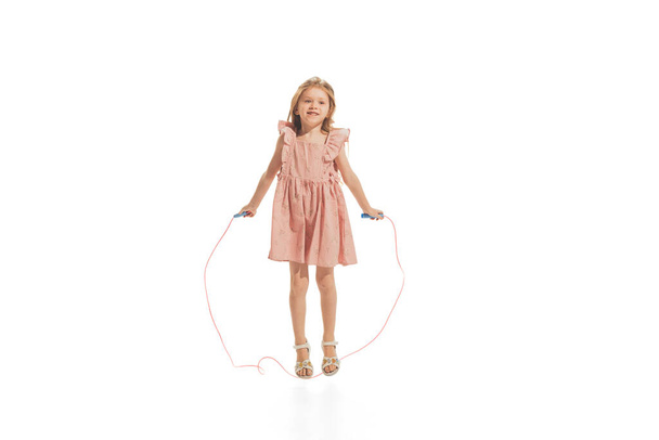 Portrait of beautiful little girl in stylish dress posing, jumping with rope, playing isolated over white studio background. Concept of childhood, game, fashion, fun, education. Copy space for ad - Foto, Imagem