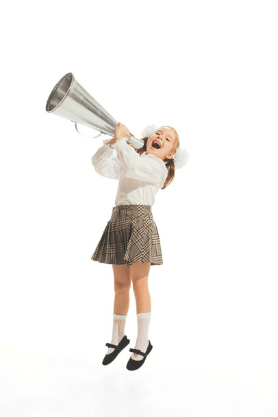 Portrait of little girl, child in school uniform with bow screaming in megaphone isolated over white studio background. Concept of childhood, emotions, game, school, fun, education. Copy space for ad - Fotografie, Obrázek