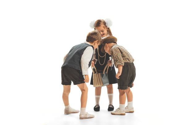 Portrait of three children, boys and girl in retro school clothes looking inside bag isolated over white studio background. Concept of childhood, game, school, fun, education. Copy space for ad - Foto, afbeelding