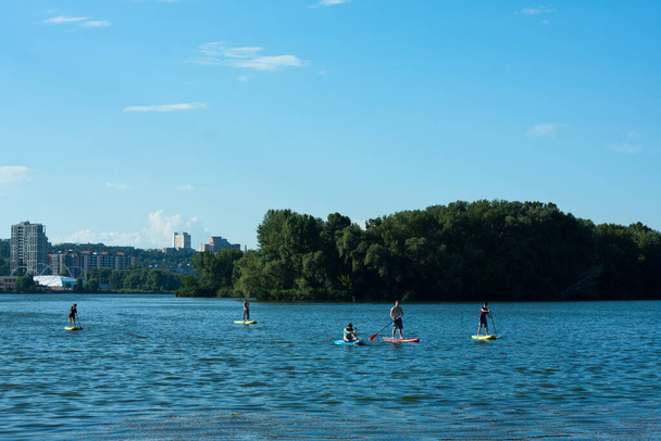 unidentified people ride SUP boards on calm river in summer on sunny day. group friends actively spend time together, do SUP on river in city. Ukraine, city Dnipro - Фото, зображення