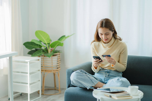Asian female consumers with credit cards and smartphones sit on their sofas at home to do banking and mobile online shopping. - Φωτογραφία, εικόνα