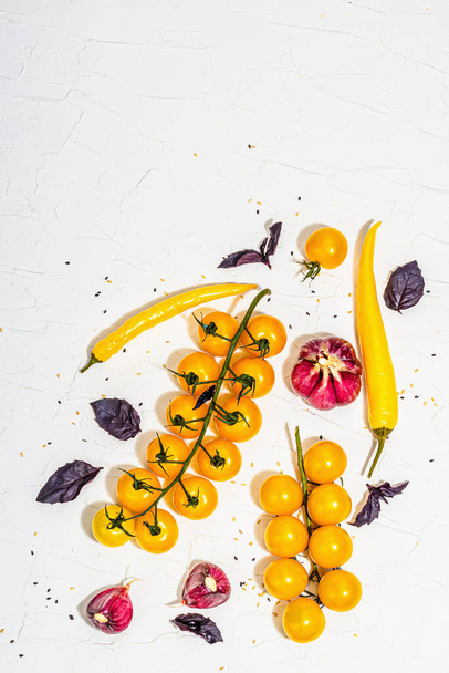 Culinary background with ripe vegetables. Yellow tomato cherries, chili pepper, garlic, purple basil. Fresh vegetables on white putty background, modern hard light, dark shadow, flat lay, top view - Photo, Image