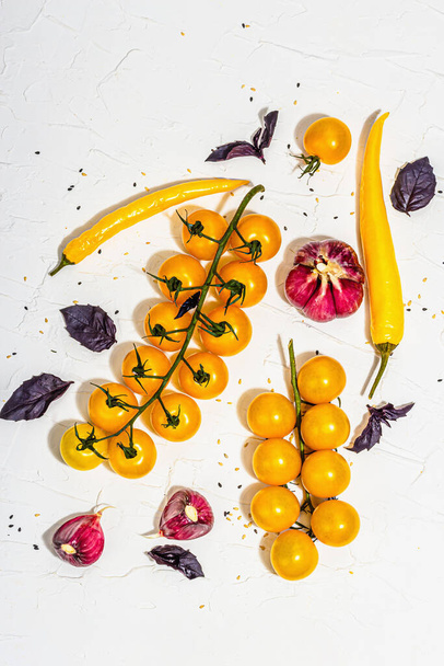 Culinary background with ripe vegetables. Yellow tomato cherries, chili pepper, garlic, purple basil. Fresh vegetables on white putty background, modern hard light, dark shadow, flat lay, top view - Photo, Image