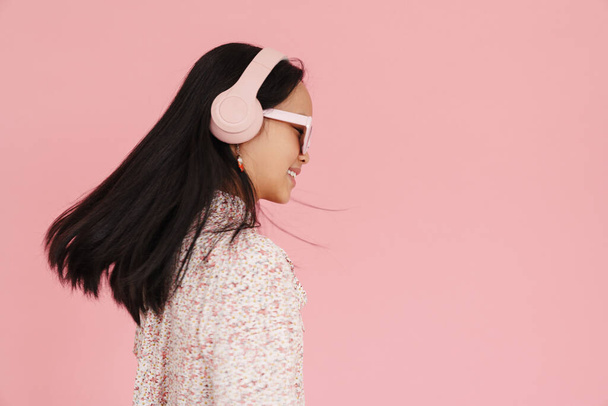 Asian girl dancing while listening music with headphones isolated over pink background - Fotó, kép