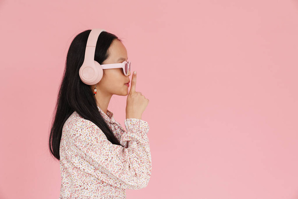 Asian girl listening music while showing silence gesture isolated over pink background - Photo, image