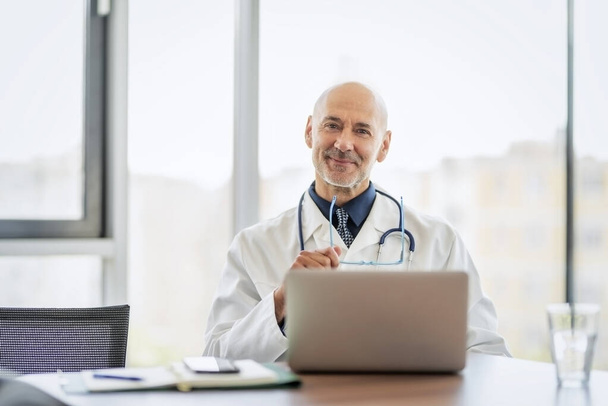 Male doctor looking at camera and smiling while sitting behind his laptop at doctors office.   - Photo, Image
