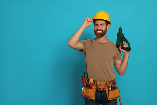 Professional builder in hard hat with tool belt and power drill on light blue background, space for text - 写真・画像