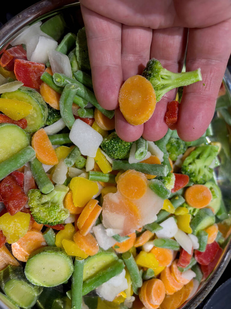 Male hand holding multiple frozen vegetables in large stainless steel pan - Foto, Imagem