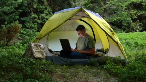 Ένας άντρας δουλεύει σε ένα λάπτοπ σε μια τουριστική κατασκήνωση σε ένα όμορφο δάσος. Έννοια της freelancing, digital nomad ή απομακρυσμένο γραφείο. 4K - Πλάνα, βίντεο
