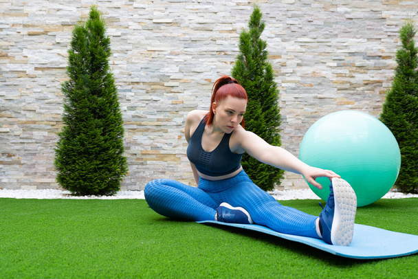 young woman with red hair, practising leg stretching exercises on a mat in the garden of her house. Sporty girl training. concept of health and well-being. natural outdoor light. - 写真・画像
