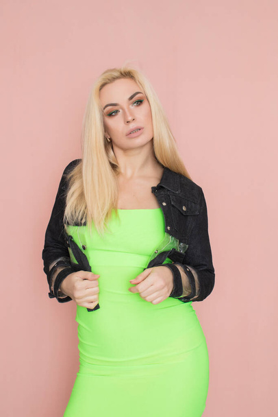 Stylish caucasian adult blonde woman wears fashionable jeans jacket and green dress, isolated over pink studio wall. - Photo, image