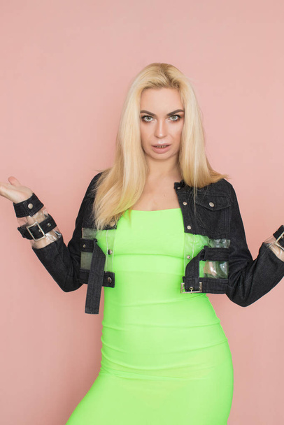 Stylish caucasian adult blonde woman wears fashionable jeans jacket and green dress, isolated over pink studio wall. - Fotografie, Obrázek