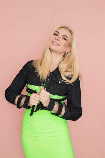 Stylish caucasian adult blonde woman wears fashionable jeans jacket and green dress, isolated over pink studio wall. - Фото, зображення