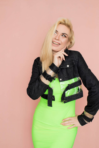 Stylish caucasian adult blonde woman wears fashionable jeans jacket and green dress, isolated over pink studio wall. - Foto, Imagem