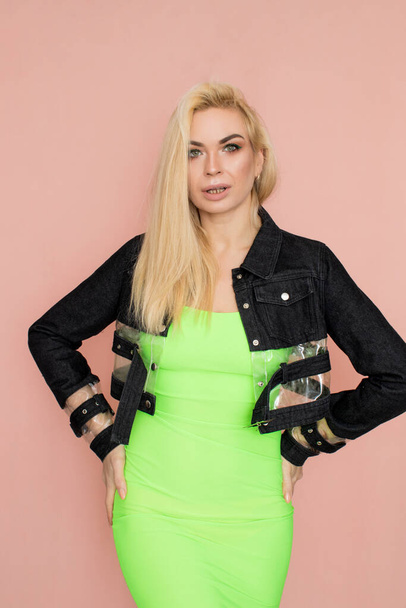 Stylish caucasian adult blonde woman wears fashionable jeans jacket and green dress, isolated over pink studio wall. - Zdjęcie, obraz