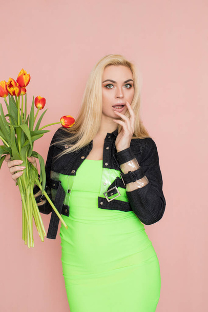 Young elegant woman in trendy jeans jacket. Blonde hair, green dress, isolated over pink background, studio shot. Fashion spring lookbook. Model woman with red tulips in her hands - Fotoğraf, Görsel
