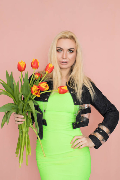 Young elegant woman in trendy jeans jacket. Blonde hair, green dress, isolated over pink background, studio shot. Fashion spring lookbook. Model woman with red tulips in her hands - Fotografie, Obrázek