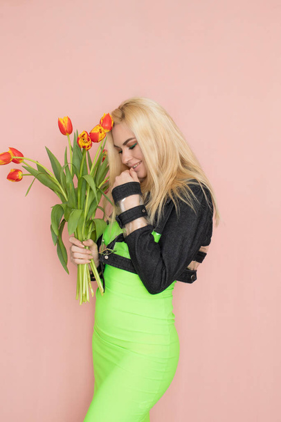 Young elegant woman in trendy jeans jacket. Blonde hair, green dress, isolated over pink background, studio shot. Fashion spring lookbook. Model woman with red tulips in her hands - Photo, Image