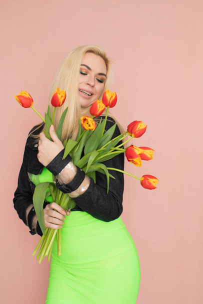 Young elegant woman in trendy jeans jacket. Blonde hair, green dress, isolated over pink background, studio shot. Fashion spring lookbook. Model woman with red tulips in her hands - Foto, immagini
