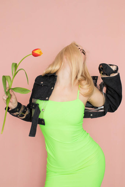 Young elegant woman in trendy jeans jacket. Blonde hair, green dress, isolated over pink background, studio shot. Fashion spring lookbook. Model woman with red tulips in her hands - Photo, image