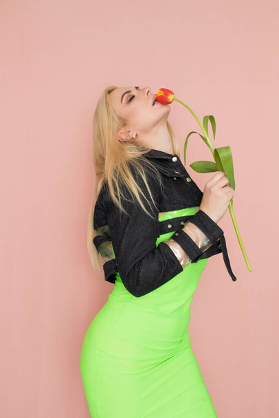 Young elegant woman in trendy jeans jacket. Blonde hair, green dress, isolated over pink background, studio shot. Fashion spring lookbook. Model woman with red tulips in her hands - Foto, Imagem