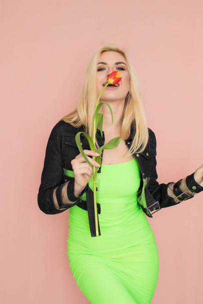 Young elegant woman in trendy jeans jacket. Blonde hair, green dress, isolated over pink background, studio shot. Fashion spring lookbook. Model woman with red tulips in her hands - Fotó, kép