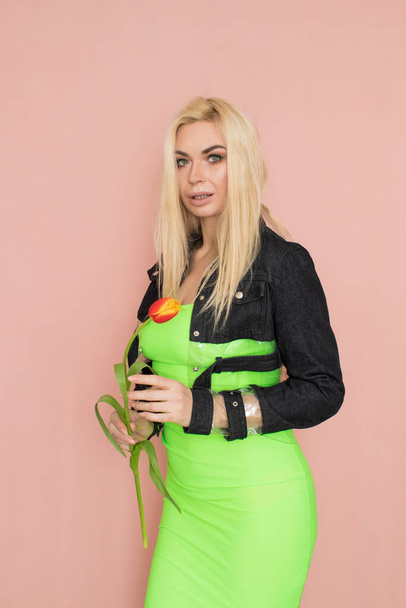 Young elegant woman in trendy jeans jacket. Blonde hair, green dress, isolated over pink background, studio shot. Fashion spring lookbook. Model woman with red tulips in her hands - Foto, immagini