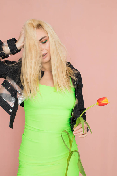 Young elegant woman in trendy jeans jacket. Blonde hair, green dress, isolated over pink background, studio shot. Fashion spring lookbook. Model woman with red tulips in her hands - Photo, image
