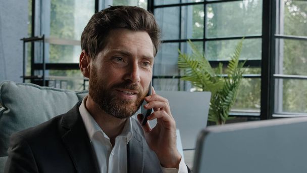 Busy bearded Caucasian adult middle-aged millennial man office worker businessman manager consulting client by telephone reading information on laptop screen looking at computer talk phone answer call - Foto, immagini
