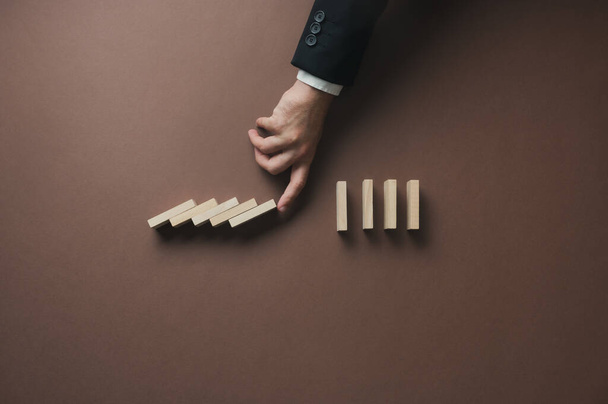 Hand of a businessman intervening a collapsing dominos to stop the disaster in a conceptual image of crisis solution. Over brown background. - Zdjęcie, obraz
